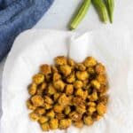 A pile of fried okra pieces on a paper towel, with two whole okra pods and a blue cloth nearby on a white surface.
