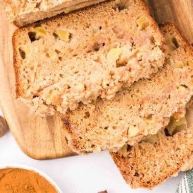 Three slices of apple bread are arranged on a wooden cutting board next to a small bowl of cinnamon, a nutmeg, and part of a striped towel.
