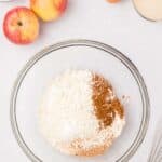 A glass bowl with flour, cinnamon, and baking powder on a white surface, surrounded by apples, an egg, sugar, apple juice, and a whisk.