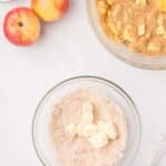 Two apples sit beside a bowl of diced butter in dry ingredients and another bowl with chunky apple batter, on a white surface.