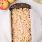 A metal loaf pan lined with parchment paper is filled with unbaked apple crumble mixture. Two whole apples and a glass bowl are nearby on a white surface.