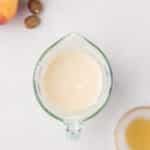 A glass measuring cup filled with a creamy liquid sits on a white surface, surrounded by apples, whole nutmeg, cinnamon sticks, and a small bowl of amber liquid.