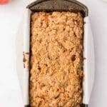A loaf pan lined with parchment paper holds a baked apple crumb bread. Surrounding the pan are apples, whole nutmeg, a cinnamon stick, and a bowl of ground cinnamon.
