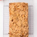 A loaf of apple streusel bread cooling on a wire rack, with apples, nutmeg, and cinnamon sticks nearby on a white surface.