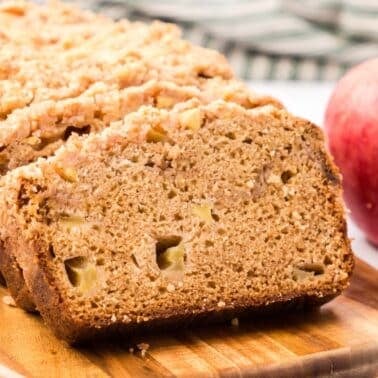 A loaf of apple bread with a crumb topping, partially sliced, sits on a wooden board next to two red apples.