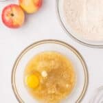 A bowl with an egg, melted butter, and liquid ingredients sits next to a bowl of flour mixture, two apples, a whisk, and a small bowl of white powder on a white surface.