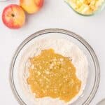 A glass bowl with flour and a wet mixture, next to diced apples, whole apples, and a small bowl of flour on a white surface.