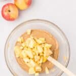 A glass bowl with light brown batter and chopped apples being mixed in, next to two whole apples, flour, and a measuring cup on a white surface.