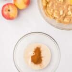 Two whole apples, a bowl of flour, a bowl of chopped apple batter, and a bowl of sugar with cinnamon on a white surface.