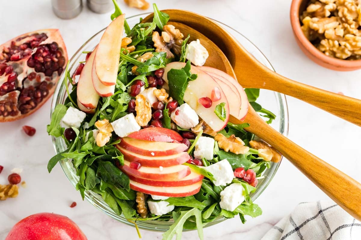 A bowl of salad with arugula, apple slices, walnuts, feta cheese, and pomegranate seeds, with wooden salad servers on top.