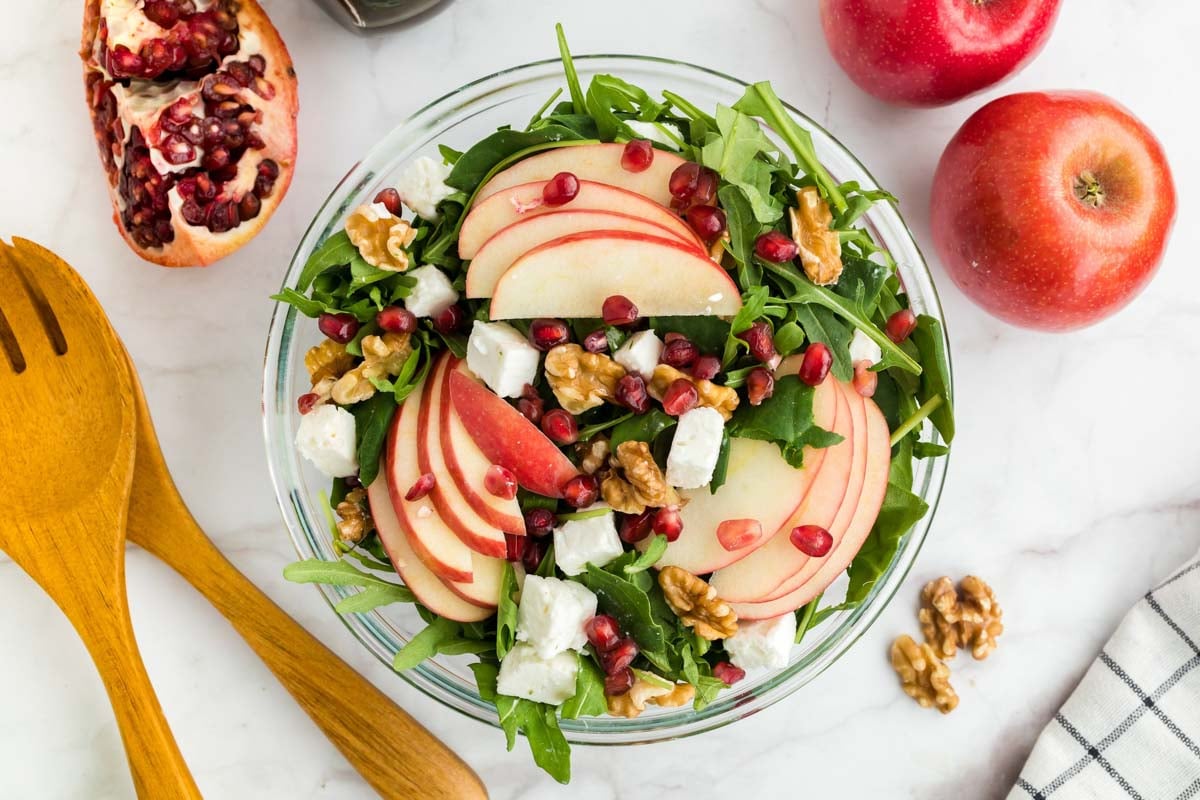 A glass bowl of salad with arugula, apple slices, feta cheese, walnuts, and pomegranate seeds, surrounded by apples, a pomegranate, and wooden salad utensils on a white surface.