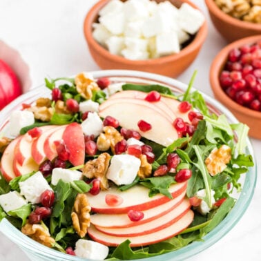A bowl of salad with arugula, sliced apples, walnuts, feta cheese, and pomegranate seeds, surrounded by bowls of walnuts, pomegranate seeds, feta, and apple slices.