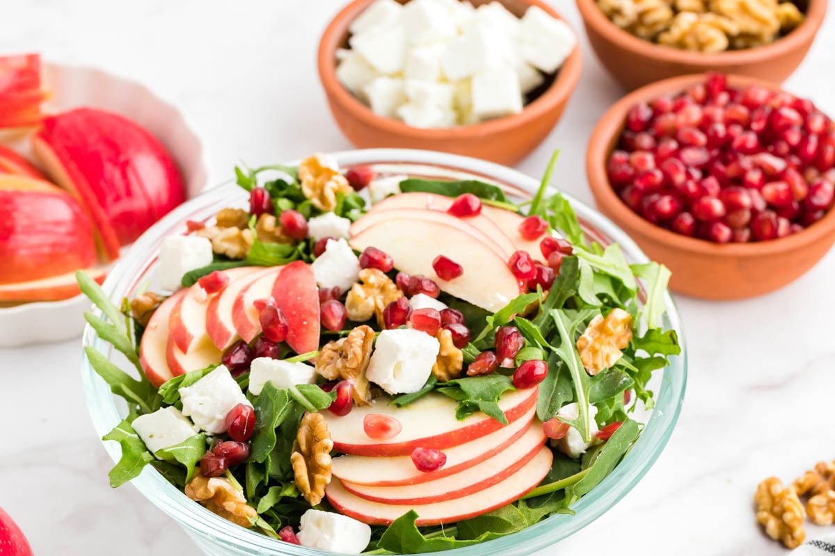 A bowl of salad with arugula, sliced apples, walnuts, feta cheese, and pomegranate seeds, surrounded by bowls of walnuts, pomegranate seeds, feta, and apple slices.