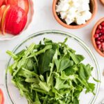 A glass bowl of fresh arugula and spinach leaves surrounded by bowls of sliced apples, feta cheese, walnuts, and pomegranate seeds.
