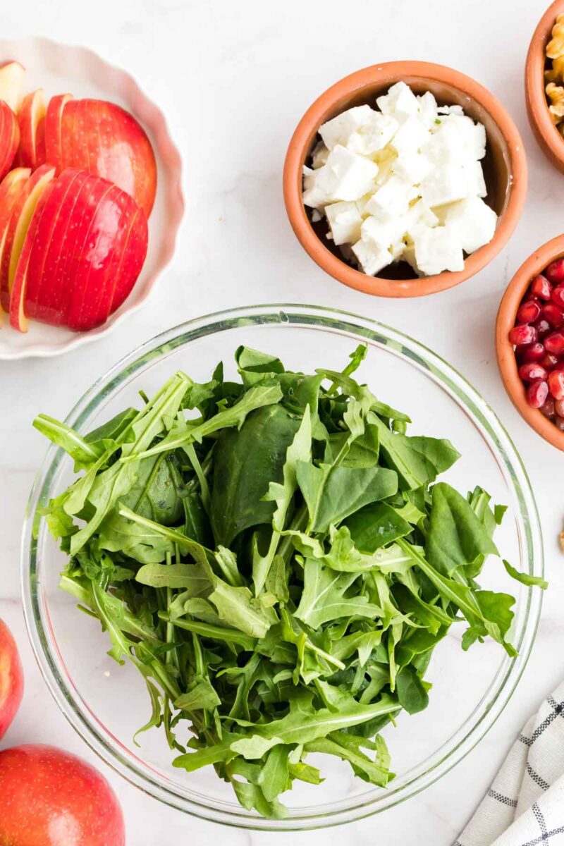 A glass bowl of fresh arugula and spinach leaves surrounded by bowls of sliced apples, feta cheese, walnuts, and pomegranate seeds.