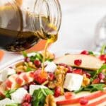 A hand pours vinaigrette dressing from a glass jar onto a salad with spinach, apple slices, walnuts, pomegranate seeds, and feta cheese in a glass bowl.