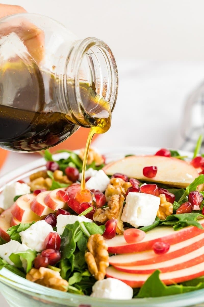 A hand pours vinaigrette dressing from a glass jar onto a salad with spinach, apple slices, walnuts, pomegranate seeds, and feta cheese in a glass bowl.
