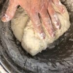 A hand covered in flour kneads a ball of dough inside a metal mixing bowl.