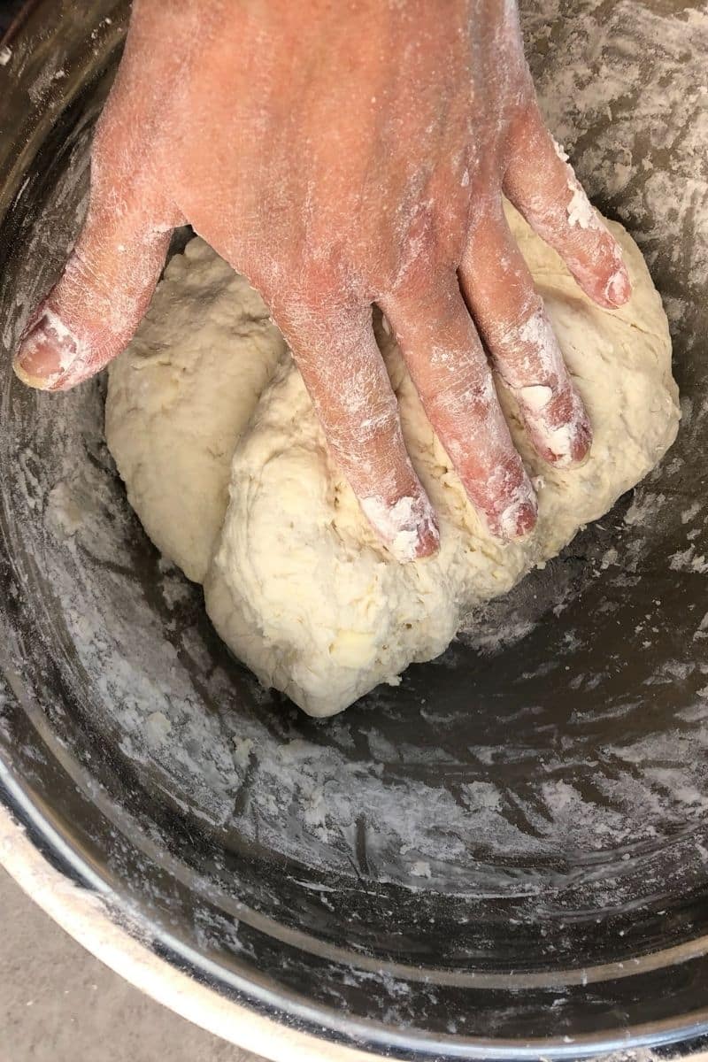 A hand covered in flour kneads a ball of dough inside a metal mixing bowl.