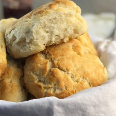 A basket lined with a white cloth filled with flaky, golden-brown biscuits, one biscuit resting on top showing its soft interior.