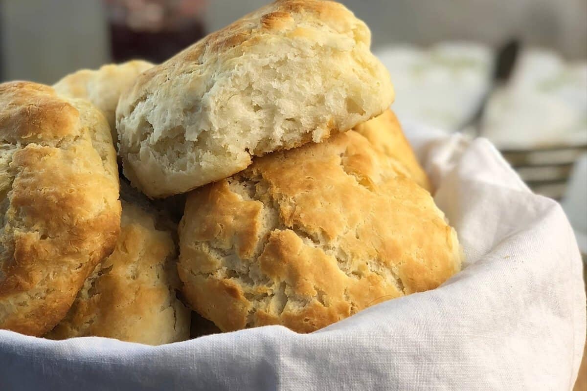 A basket lined with a white cloth filled with flaky, golden-brown biscuits, one biscuit resting on top showing its soft interior.