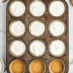 A muffin tin with twelve cups, some filled with white batter and others with a crumb crust. A spoon, cinnamon sticks, and an apple are nearby on a marble surface.