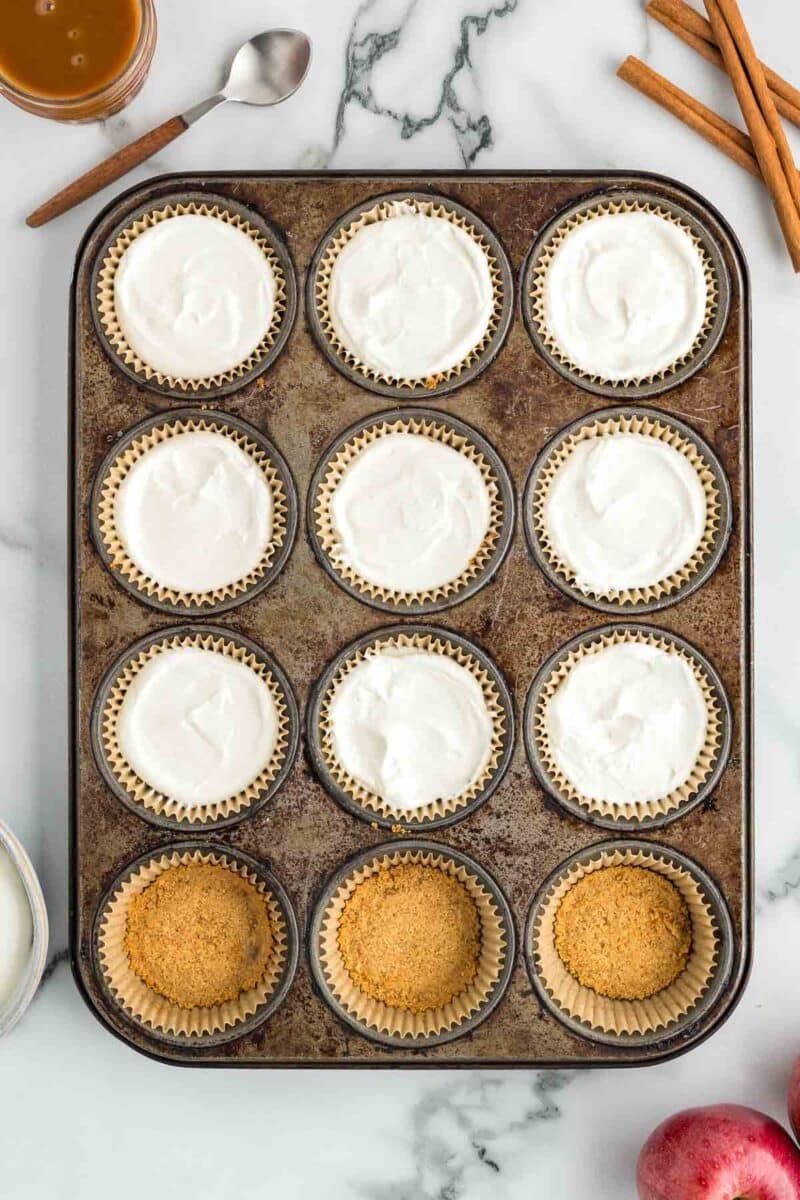 A muffin tin with twelve cups, some filled with white batter and others with a crumb crust. A spoon, cinnamon sticks, and an apple are nearby on a marble surface.