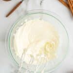A glass bowl of whipped cream cheese mixture sits on a marble countertop with a hand mixer, caramel sauce, cinnamon sticks, and apples nearby.