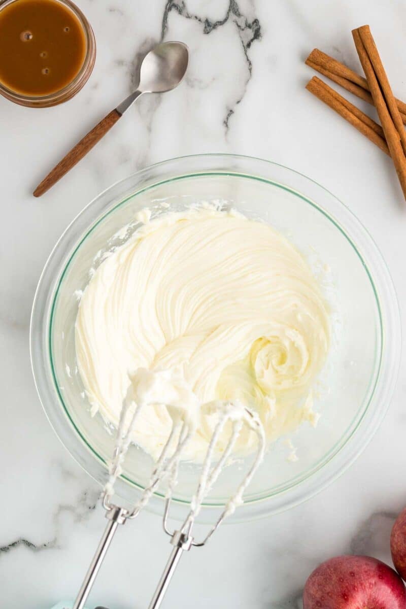 A glass bowl of whipped cream cheese mixture sits on a marble countertop with a hand mixer, caramel sauce, cinnamon sticks, and apples nearby.