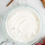 A glass bowl of whipped cream with electric beaters, surrounded by apples, cinnamon sticks, a spoon, and a small jar of caramel on a marble surface.