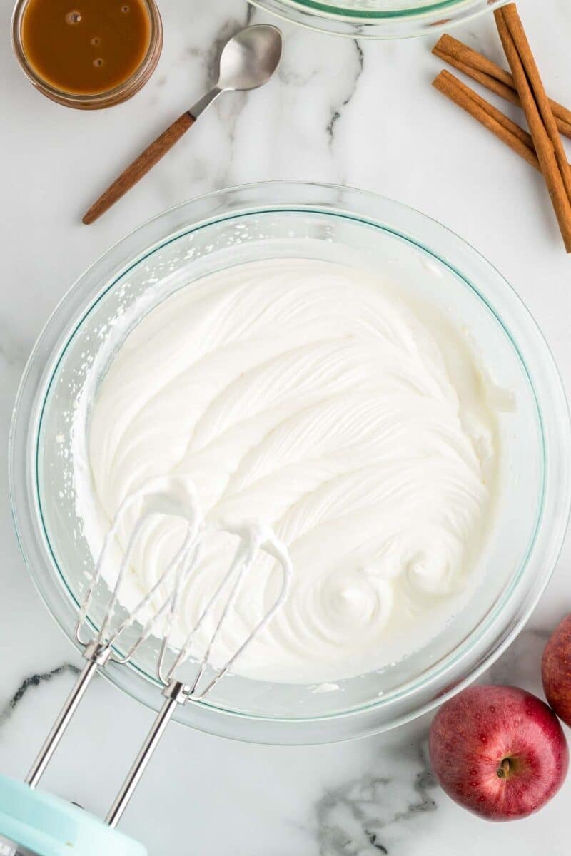 A glass bowl of whipped cream with electric beaters, surrounded by apples, cinnamon sticks, a spoon, and a small jar of caramel on a marble surface.