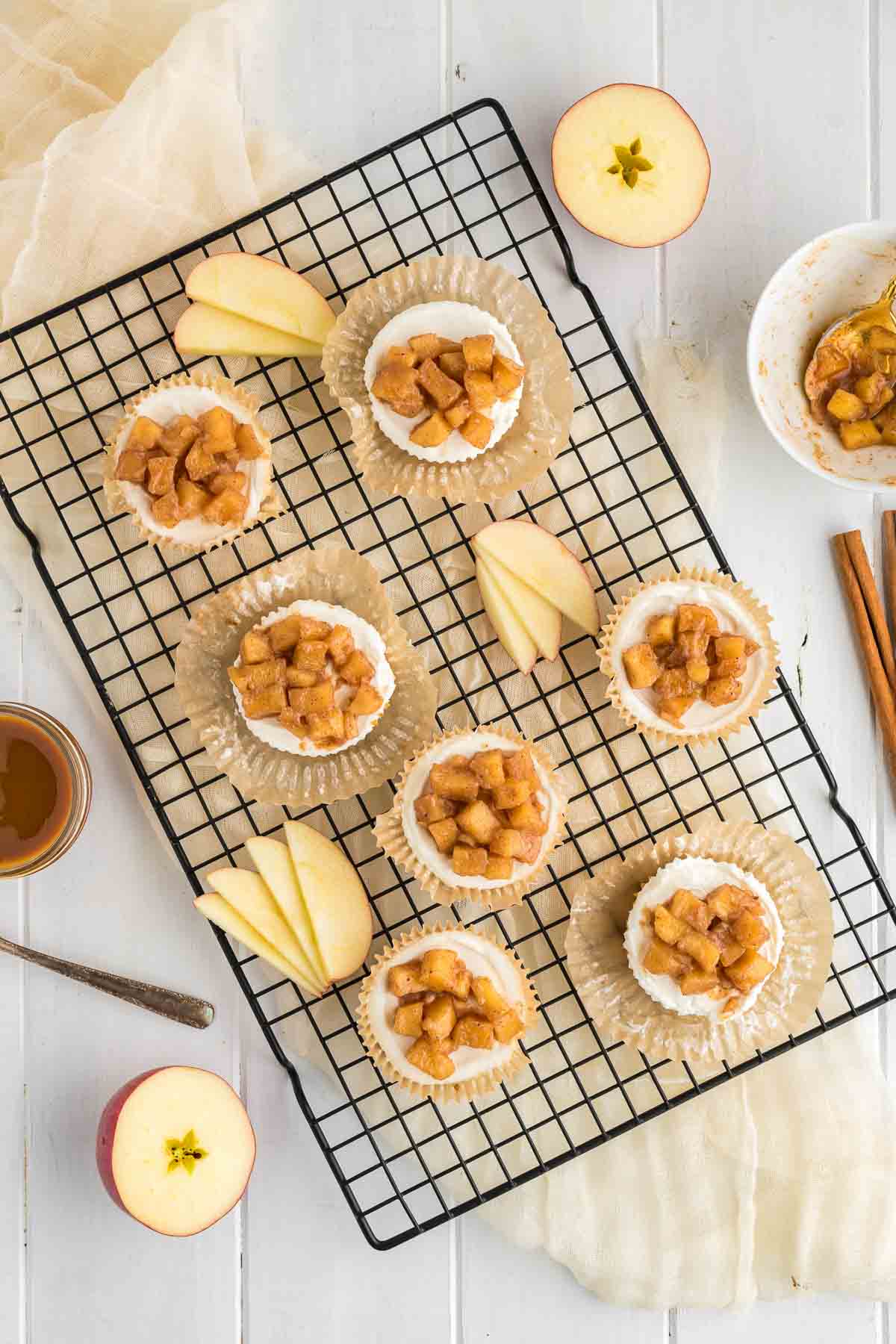 Mini cheesecakes topped with diced apple mixture on a cooling rack, surrounded by apple slices, whole apples, cinnamon sticks, and a small bowl of topping.