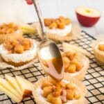A hand drizzles caramel sauce from a spoon onto a mini cheesecake topped with diced apples on a cooling rack. Sliced apples and more desserts are in the background.