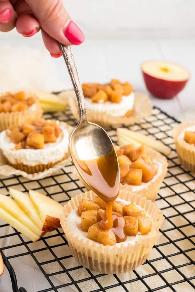 A hand drizzles caramel sauce from a spoon onto a mini cheesecake topped with diced apples on a cooling rack. Sliced apples and more desserts are in the background.