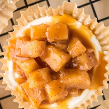 A close-up of a mini caramel apple cheesecake with diced caramelized apples and sauce on a creamy base in a paper cup, set on a cooling rack.