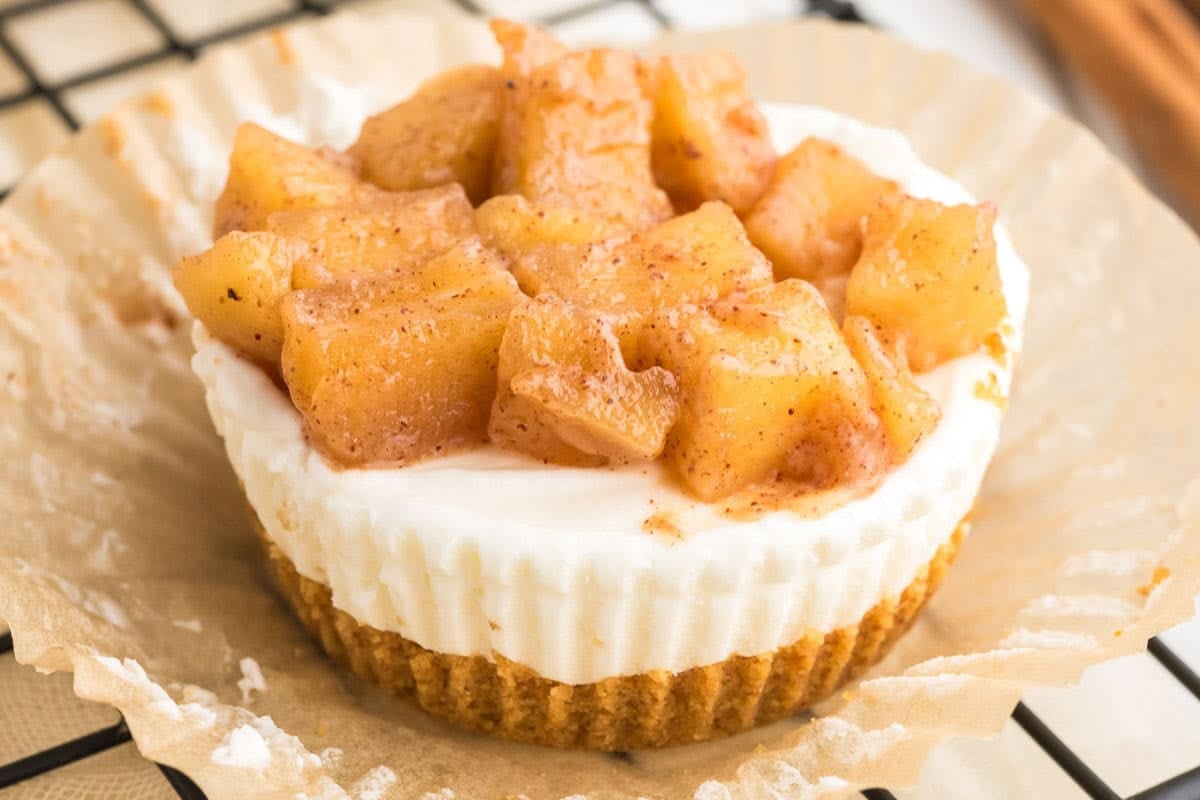 A mini caramel apple cheesecake topped with cinnamon-spiced apple chunks sits on a crinkled brown paper liner.