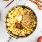 A saucepan with chopped apples, a chunk of butter, brown sugar, and spices, photographed on a marble surface with apples, cinnamon sticks, a spoon, and caramel sauce nearby.