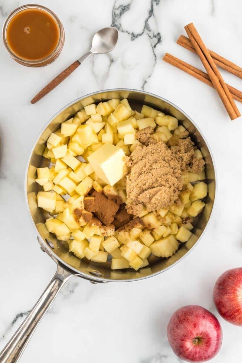 A saucepan with chopped apples, a chunk of butter, brown sugar, and spices, photographed on a marble surface with apples, cinnamon sticks, a spoon, and caramel sauce nearby.