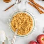 A bowl of crumbly mixture with a spoon, surrounded by apples, cinnamon sticks, a glass of caramel, baking cups, and baking utensils on a marble surface.