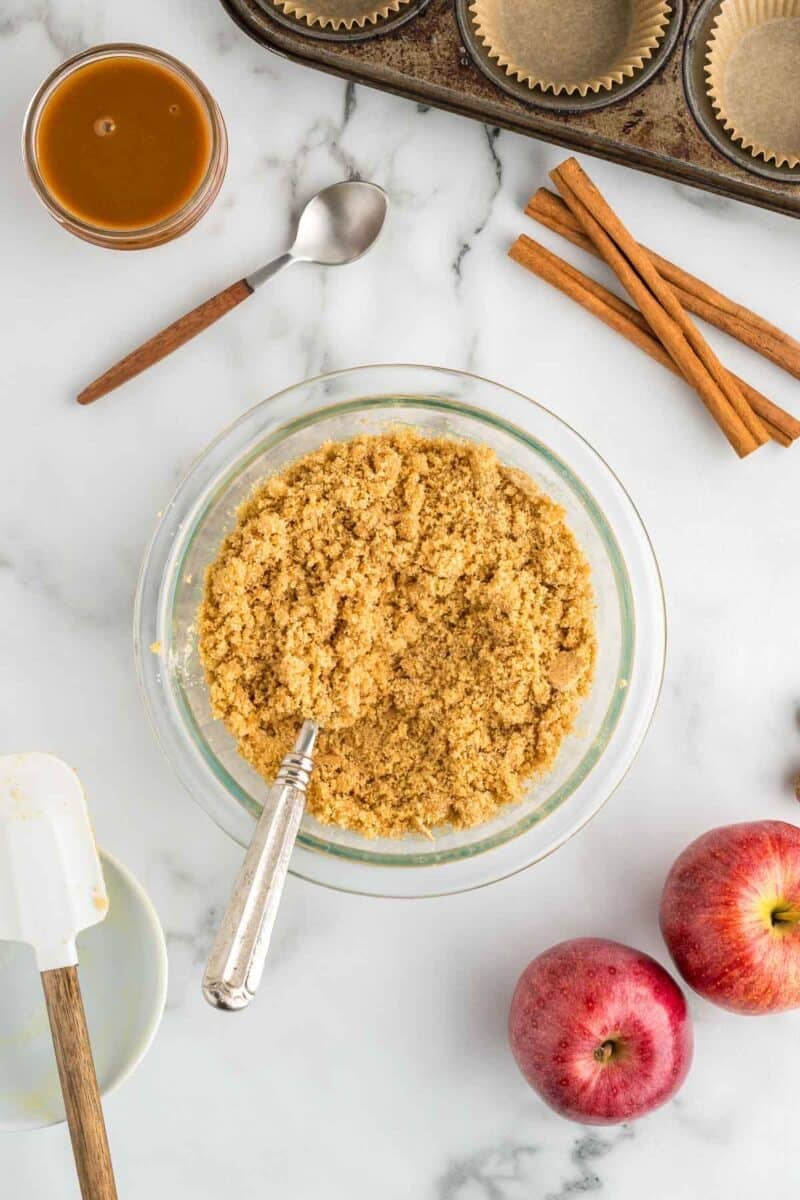 A bowl of crumbly mixture with a spoon, surrounded by apples, cinnamon sticks, a glass of caramel, baking cups, and baking utensils on a marble surface.