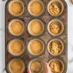 A hand presses crumb mixture into a muffin tin lined with paper cups, while some cups are partially filled; cinnamon sticks, a spoon, and brown sugar are nearby.