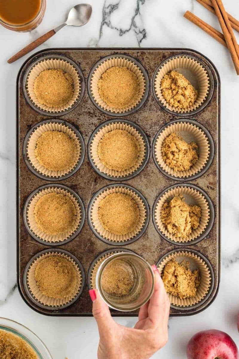 A hand presses crumb mixture into a muffin tin lined with paper cups, while some cups are partially filled; cinnamon sticks, a spoon, and brown sugar are nearby.