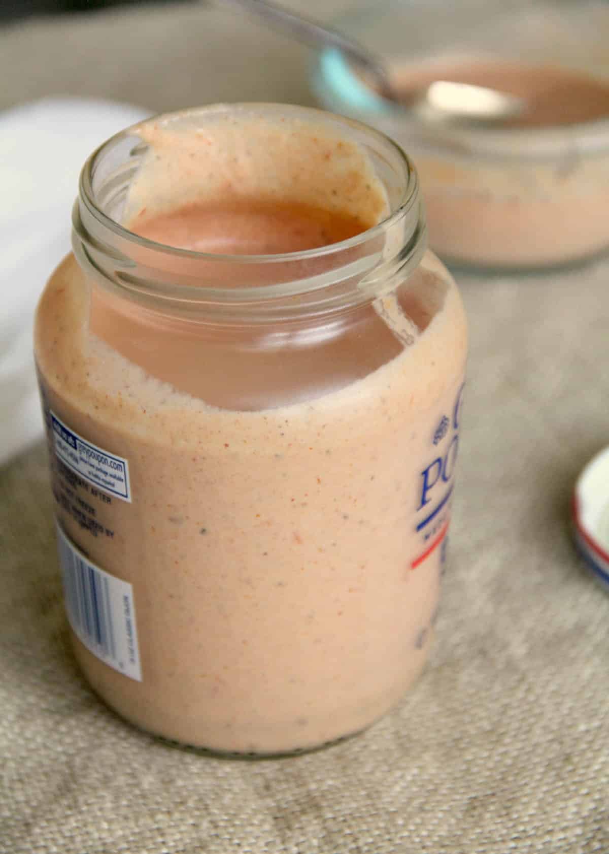 An open glass jar filled with a creamy, light orange sauce sits on a beige surface, with a spoon and another container blurred in the background.