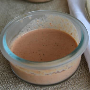 A small glass bowl containing a creamy, orange-colored sauce sits on a textured surface next to a white napkin and a metal spoon.