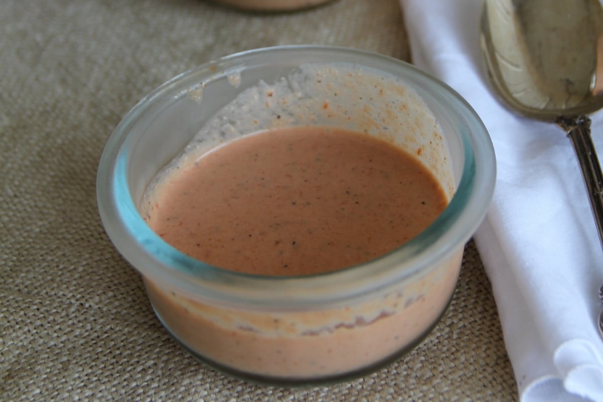 A small glass bowl containing a creamy, orange-colored sauce sits on a textured surface next to a white napkin and a metal spoon.
