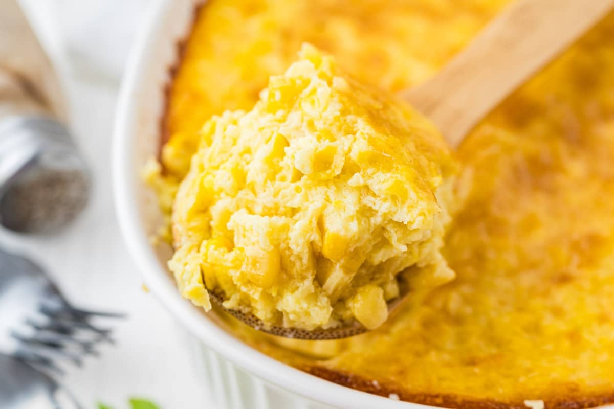 A wooden spoon holds a serving of baked corn casserole, showing its creamy, golden texture, with the rest of the casserole in a white dish in the background.