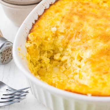 Baked corn casserole in a white dish with a portion missing, next to two forks, stacked bowls, and a salt shaker on a white surface.