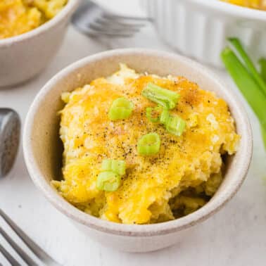 A bowl of cheesy casserole topped with chopped green onions and black pepper, with a fork, green onions, and a pepper shaker nearby.
