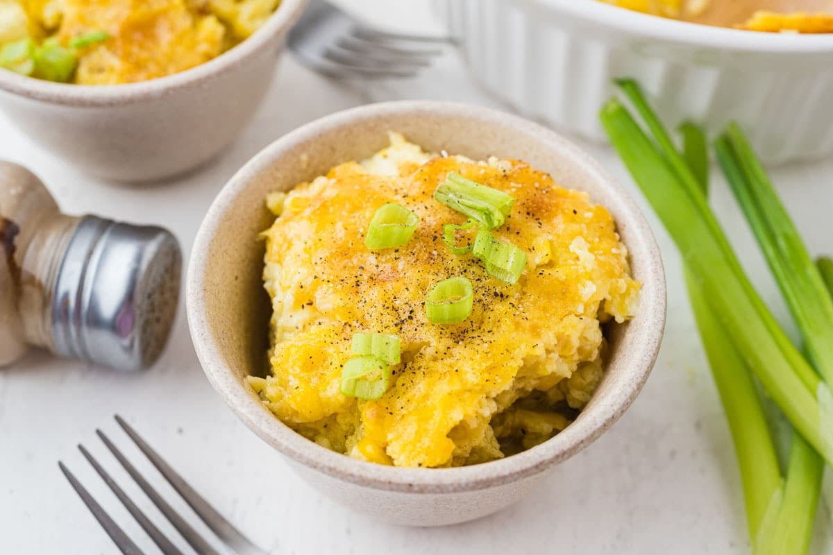 A bowl of cheesy casserole topped with chopped green onions and black pepper, with a fork, green onions, and a pepper shaker nearby.