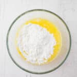 A glass bowl containing melted butter and a mound of white powdered sugar on a white surface.